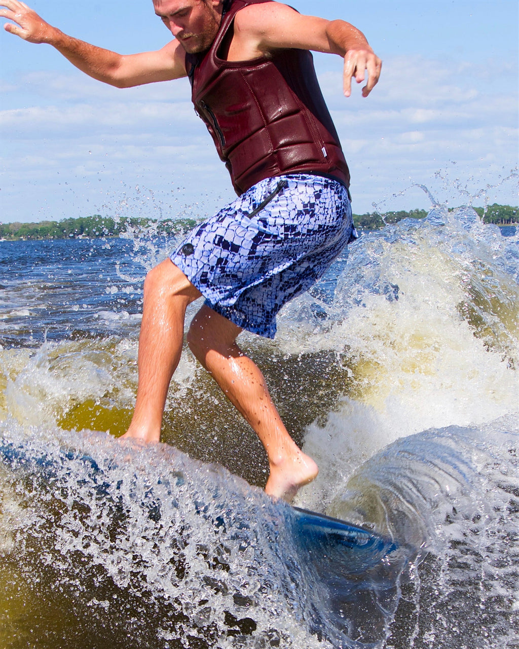A man surfing on a wave in the water, wearing a dark red life vest and blue patterned swim trunks, with white spray and a blue sky in the background.