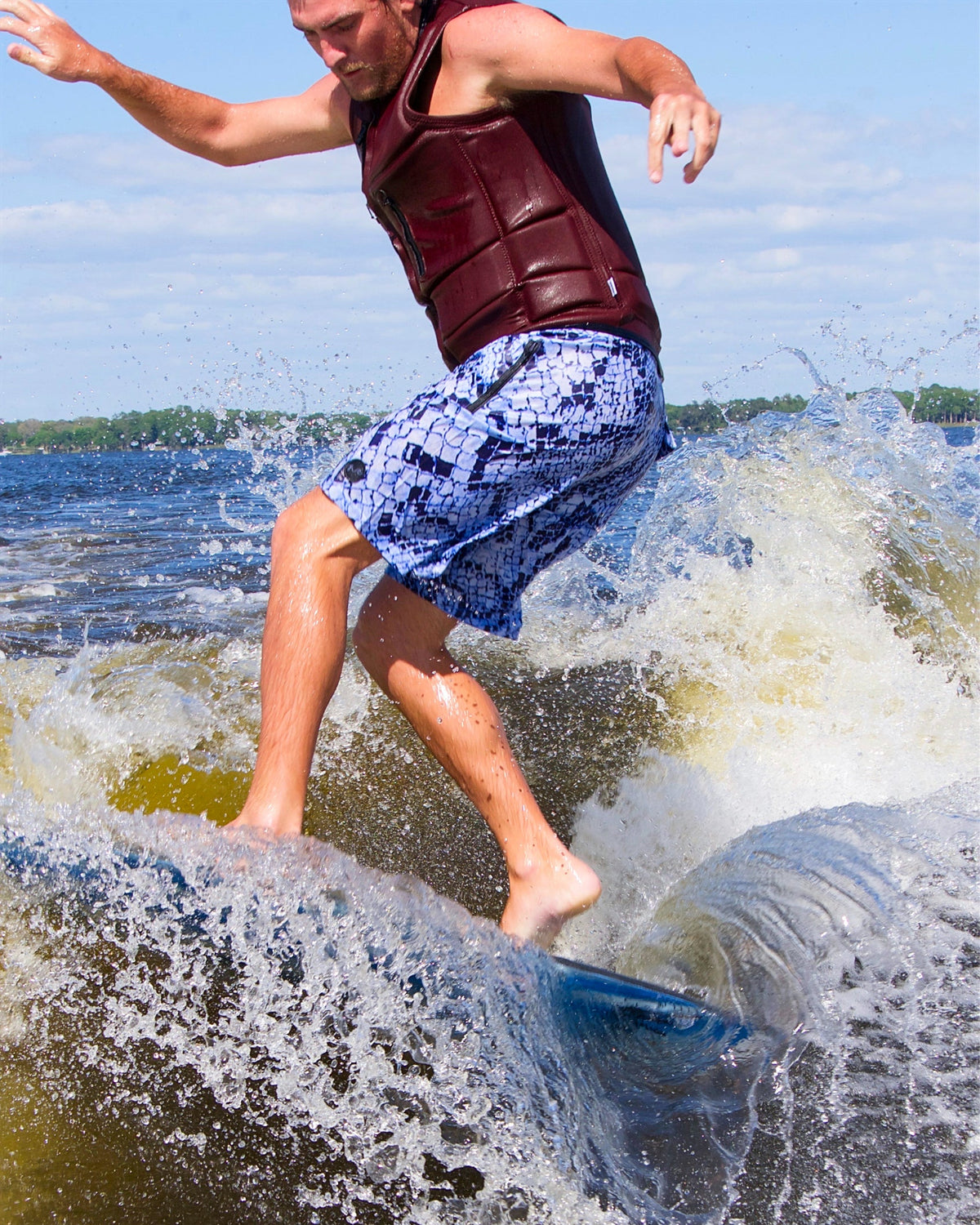 A man surfing on a wave in the water, wearing a dark red life vest and blue patterned swim trunks, with white spray and a blue sky in the background.