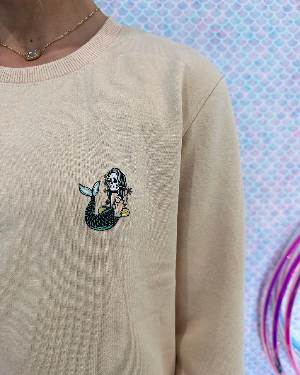 A close-up of a person wearing a beige crewneck sweatshirt with a small embroidered mermaid graphic on the left chest. The background is a sparkly, iridescent pink and blue surface.