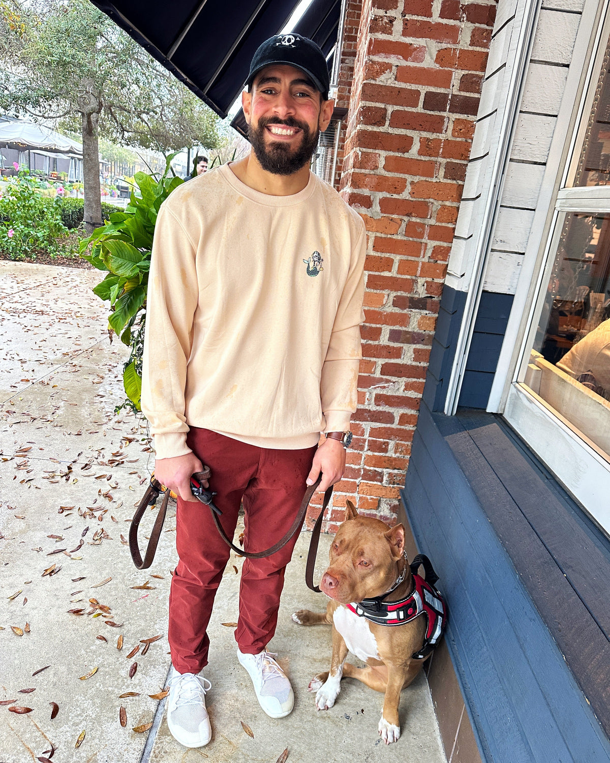 A man wearing a beige crewneck sweatshirt, rust-colored pants, and white sneakers stands on a sidewalk next to a brick building, holding a leash with a brown and white pitbull-type dog wearing a red harness.