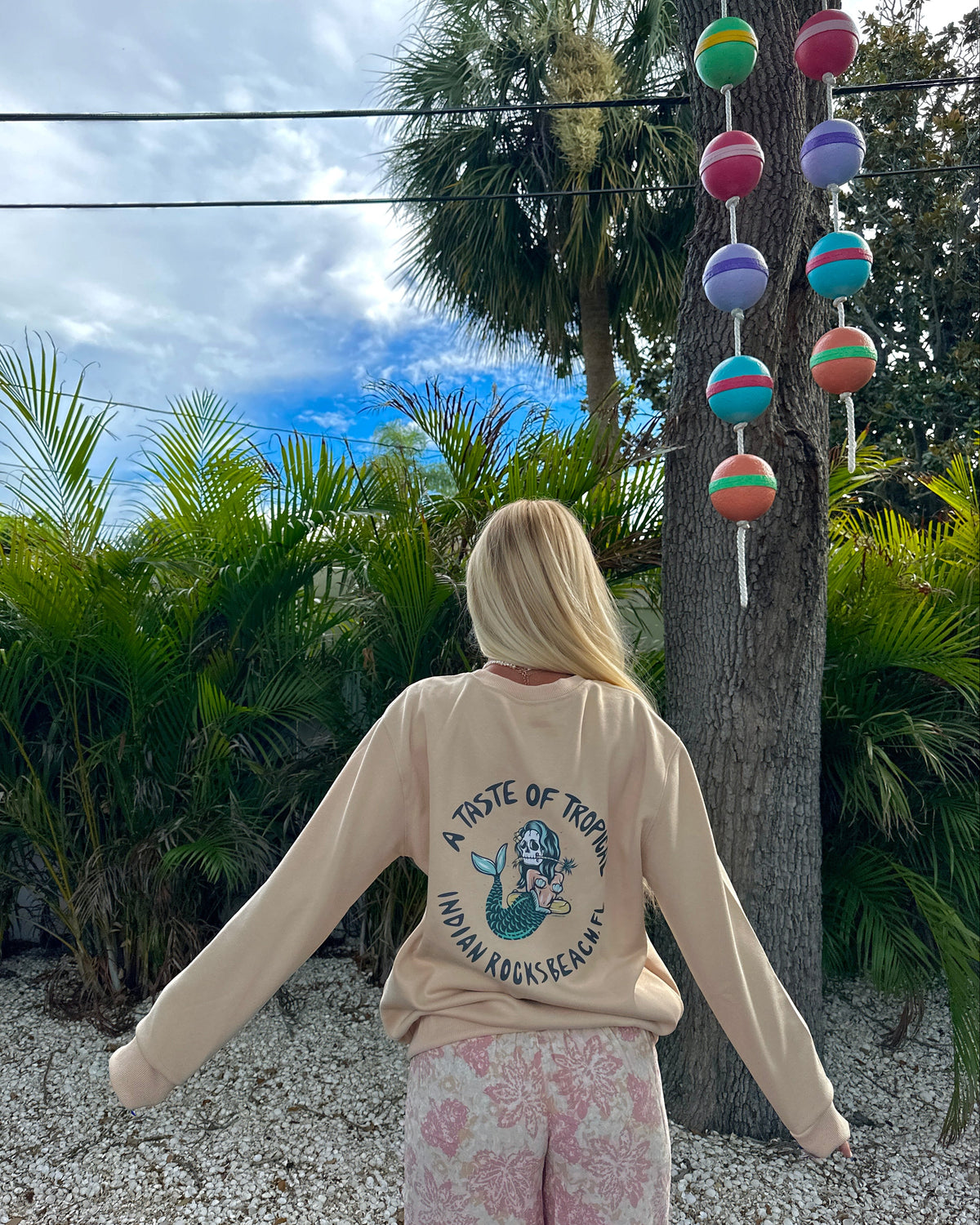 A woman stands outdoors facing a tree, wearing a beige sweatshirt with a mermaid graphic and the text 'A PRIDE OF TROPICAL PINON KNOCKHEADS' and pink floral pants. Colorful hanging marbles are visible on the tree.