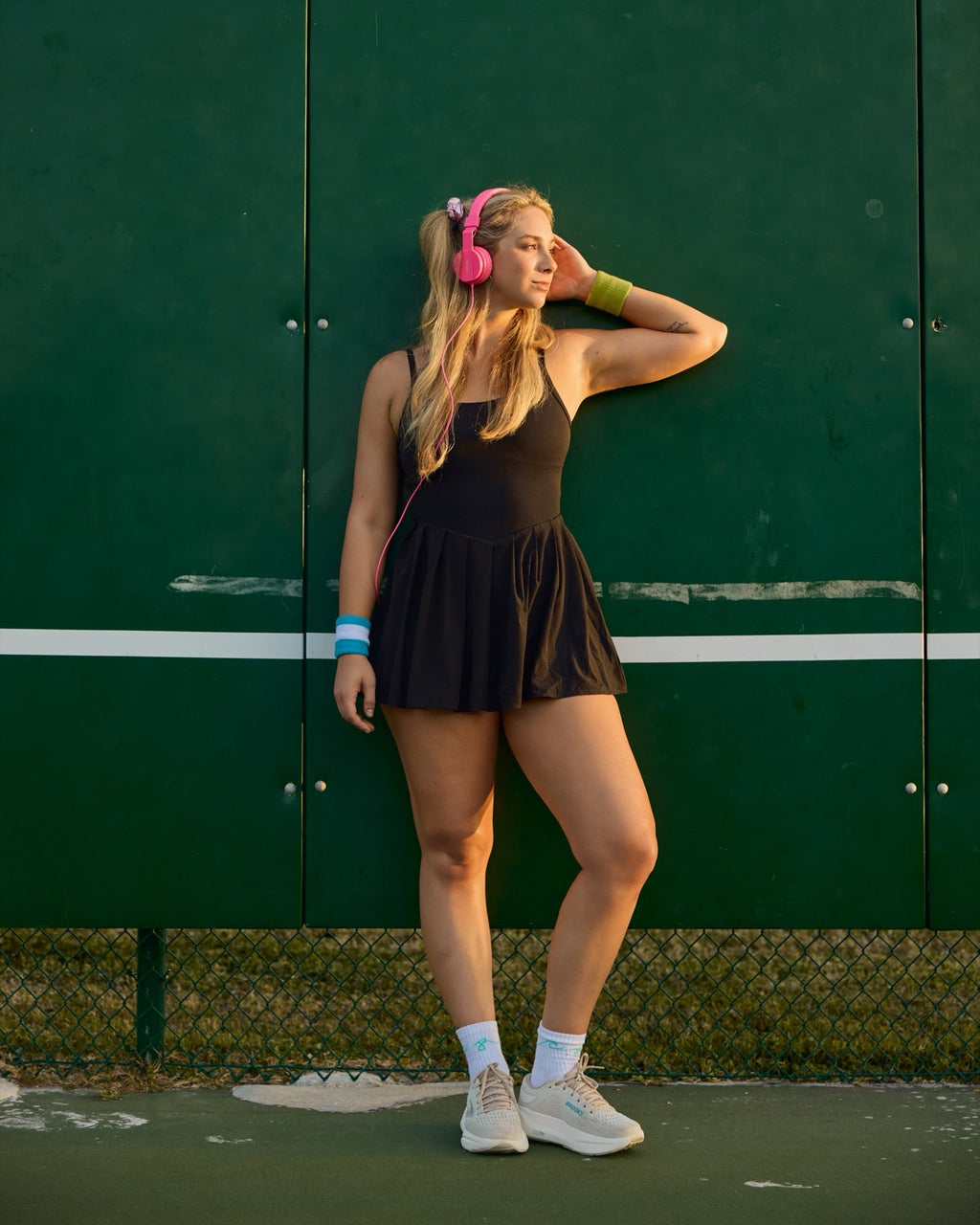 A woman posing on a tennis court against a dark green wall, wearing a black tennis dress, pink headphones, and white sneakers.