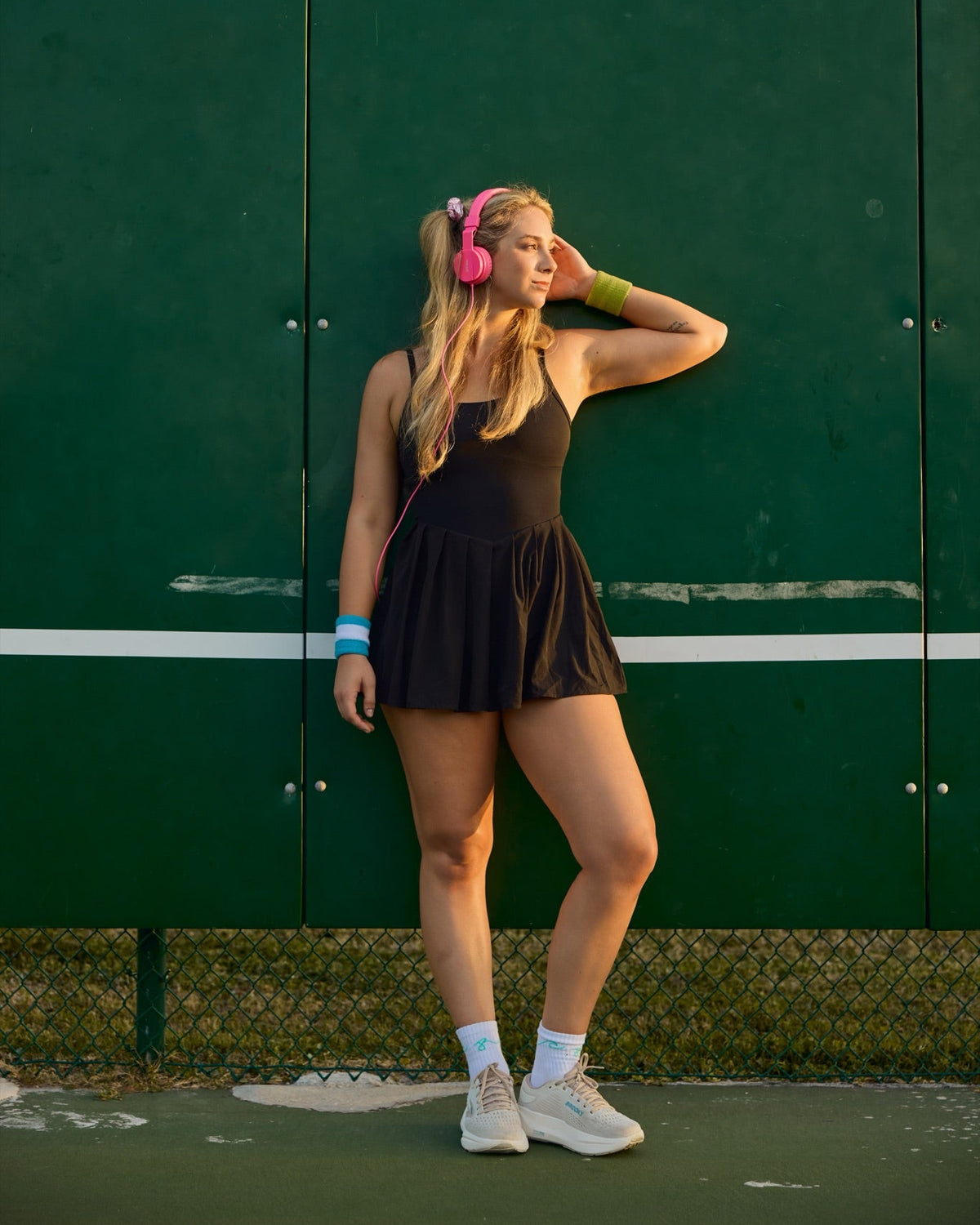 A woman posing on a tennis court against a dark green wall, wearing a black tennis dress, pink headphones, and white sneakers.