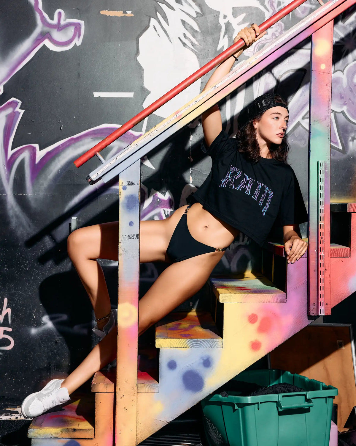 A woman posing on graffiti-covered outdoor stairs wearing a black graphic t-shirt with 'KRANTI' printed on the front, black bikini bottoms, and white sneakers.
