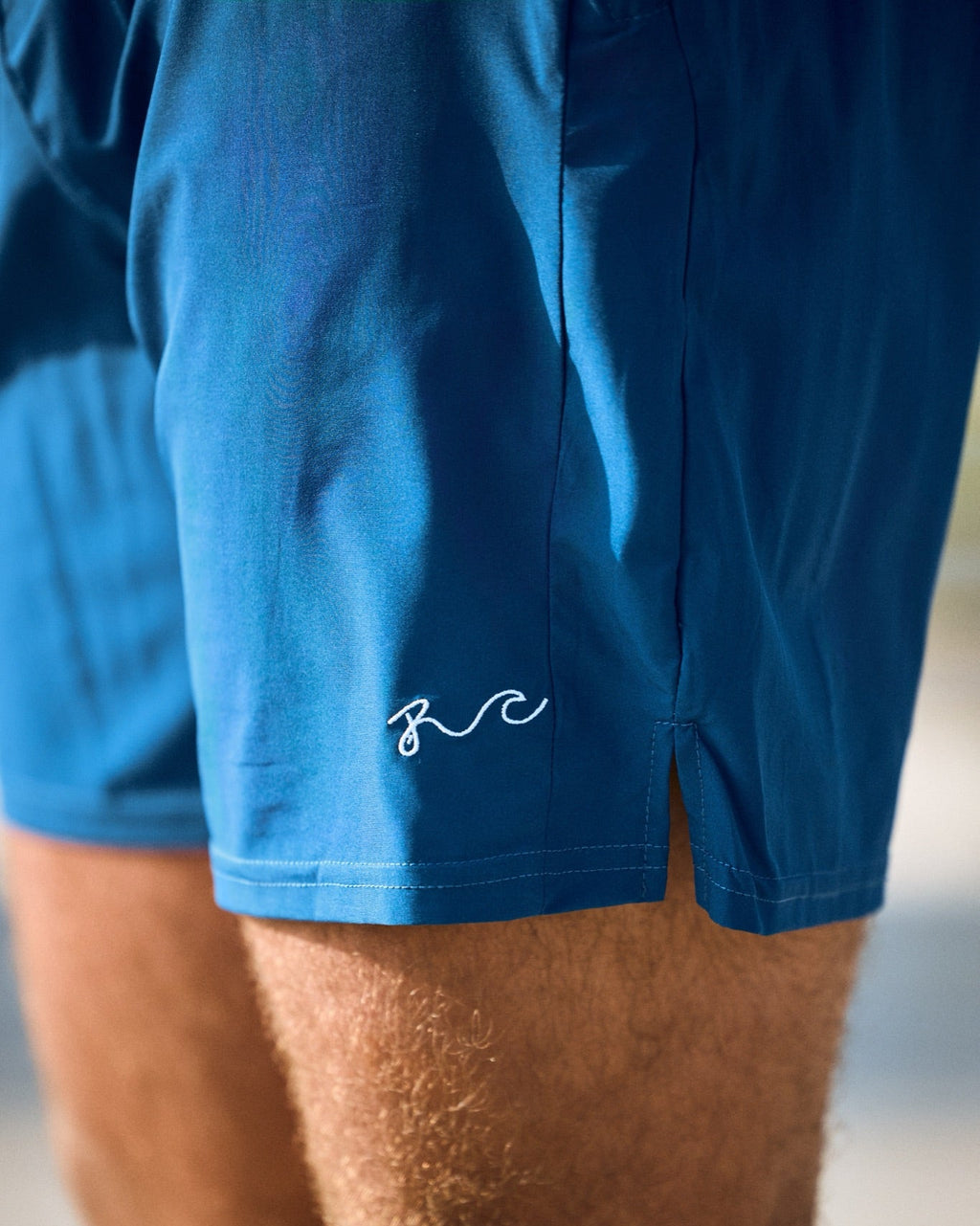 Close-up of a person wearing blue athletic shorts with a small white script logo near the hem, showing the legs against a softly blurred light background.