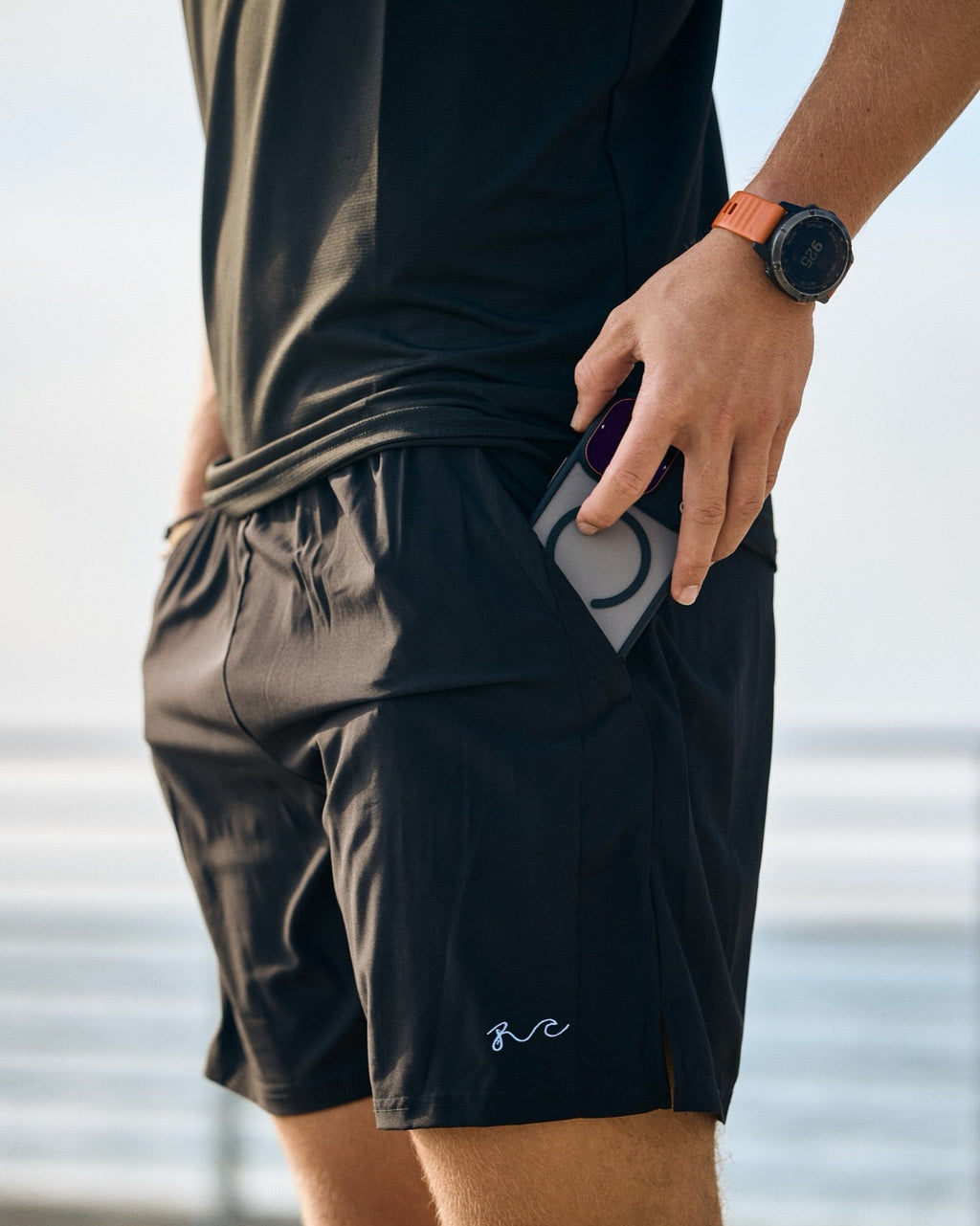 A close-up of a person wearing black athletic shorts with a side pocket holding a phone, outdoors near the water. The shorts have a small white script logo near the lower left leg.