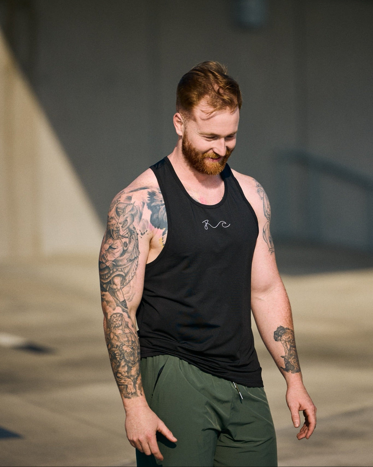 A man with tattoos and a beard wearing a black athletic tank top with a small white script logo on the chest, paired with olive green sweatpants, walking outdoors under natural sunlight.