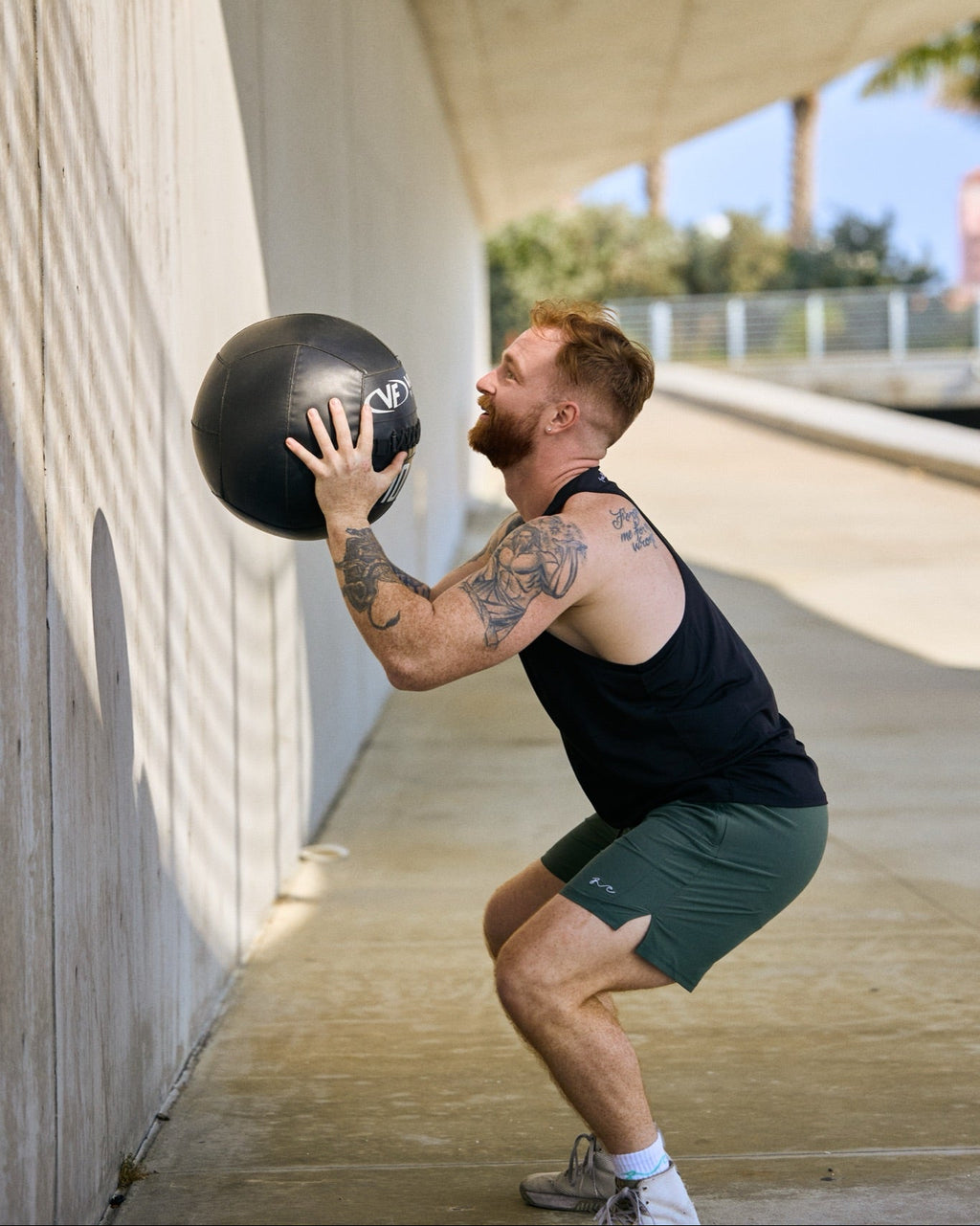 A man in a black tank top and green athletic shorts squats while holding a black medicine ball against a concrete wall in an outdoor covered walkway.