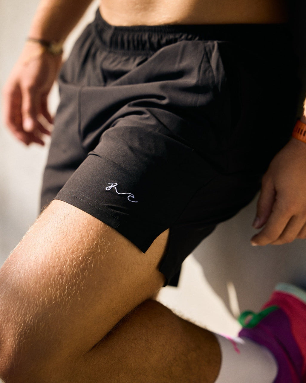 Close-up of a person wearing black athletic shorts with a small white embroidered logo near the left hem, shown against a light neutral background.