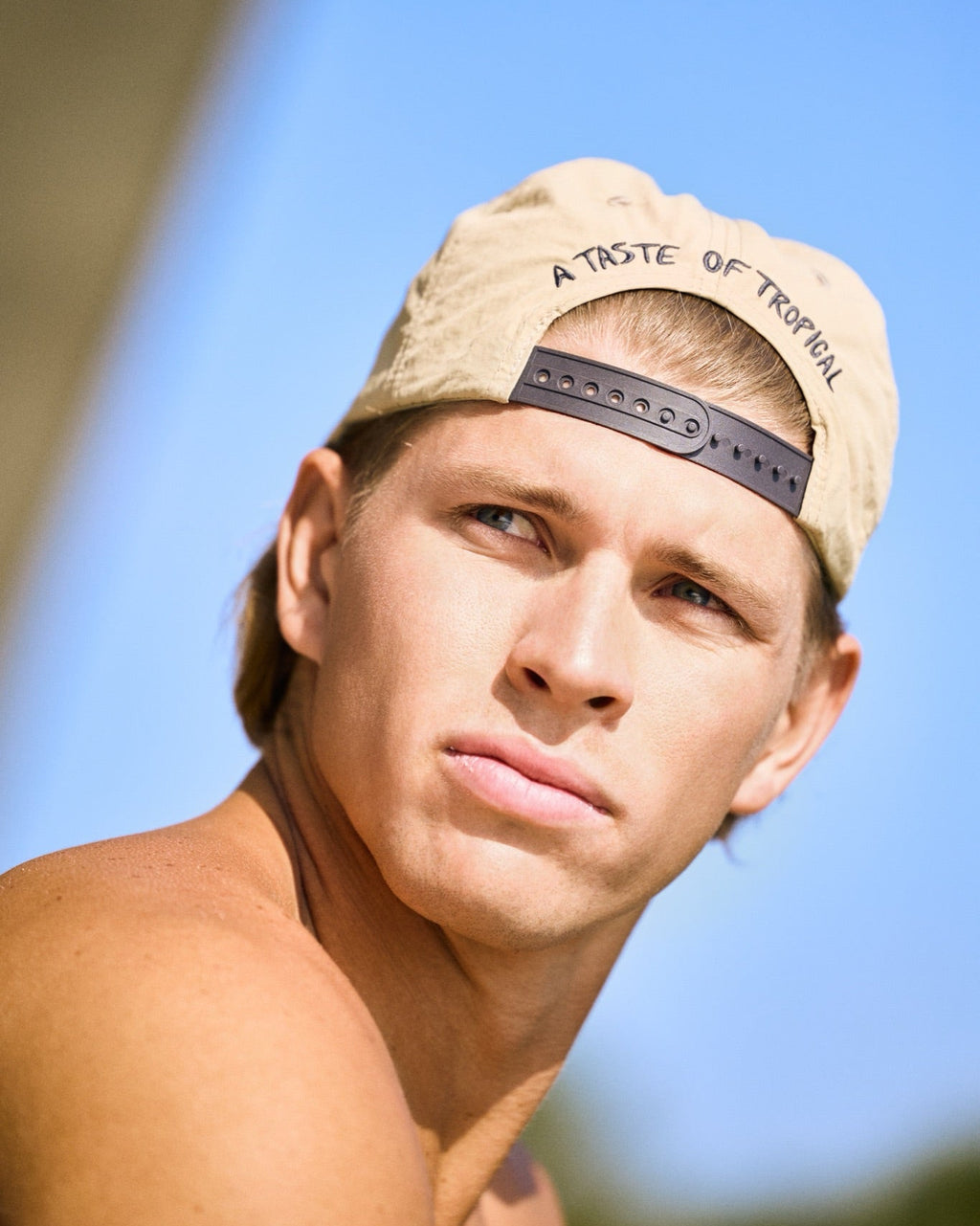 A close-up shot of a man wearing a beige baseball cap backwards with the text 'A TASTE OF TROPICAL' printed on it, set against a bright blue sky background.