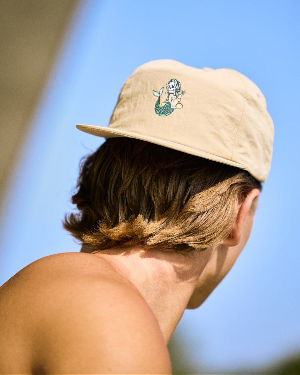 A man shown from behind wearing a beige baseball cap with a mermaid graphic on the front, set against a bright blue sky.