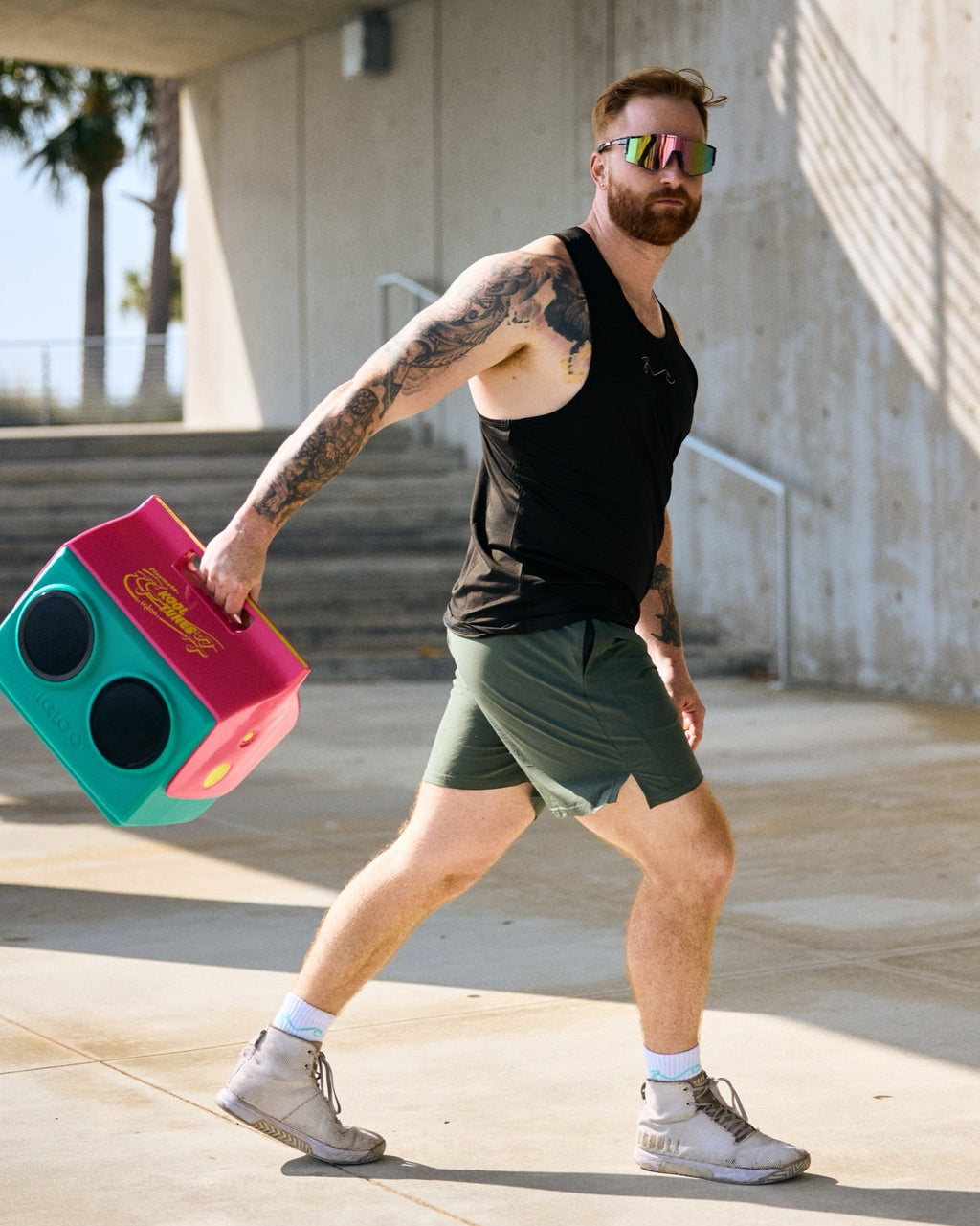 A man wearing a black tank top, green shorts, and sunglasses walks while carrying a colorful pink and teal foam exercise cube in an outdoor concrete walkway.