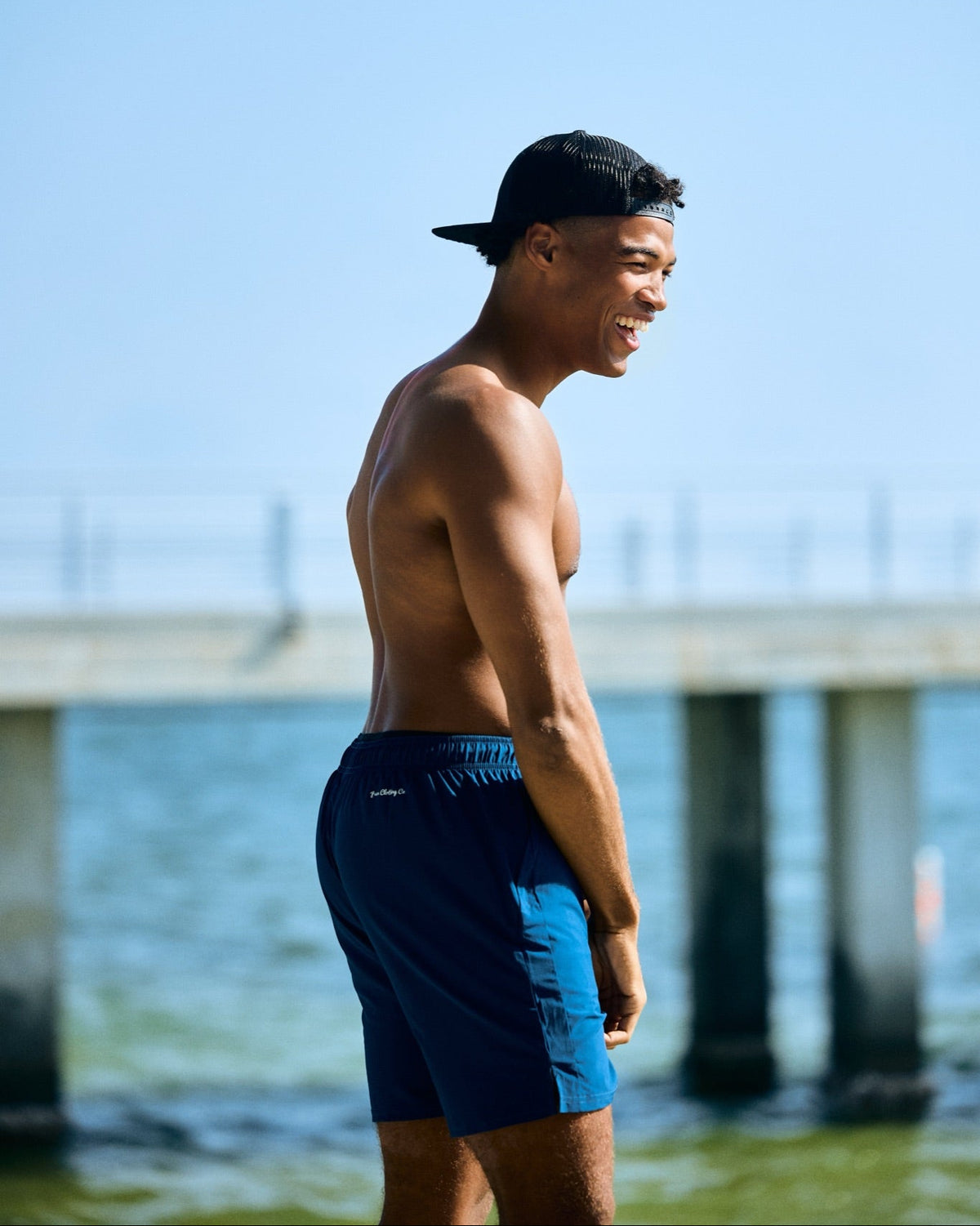 A shirtless man standing outdoors by the water near a pier, wearing blue swim shorts and a black baseball cap.
