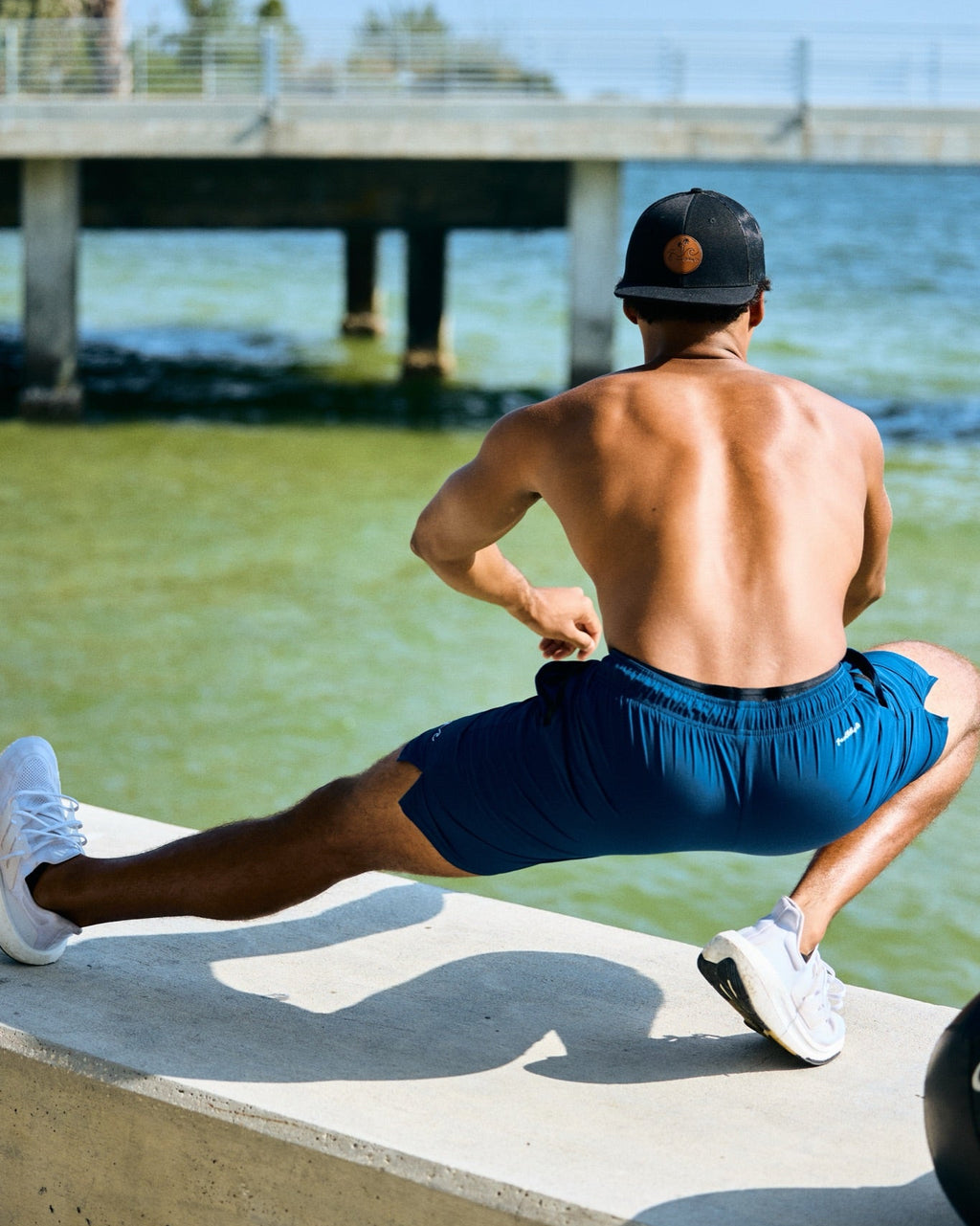 A shirtless man wearing blue athletic shorts, white sneakers, and a black baseball cap stretches on a concrete pier next to the ocean, with a bridge and water in the background.