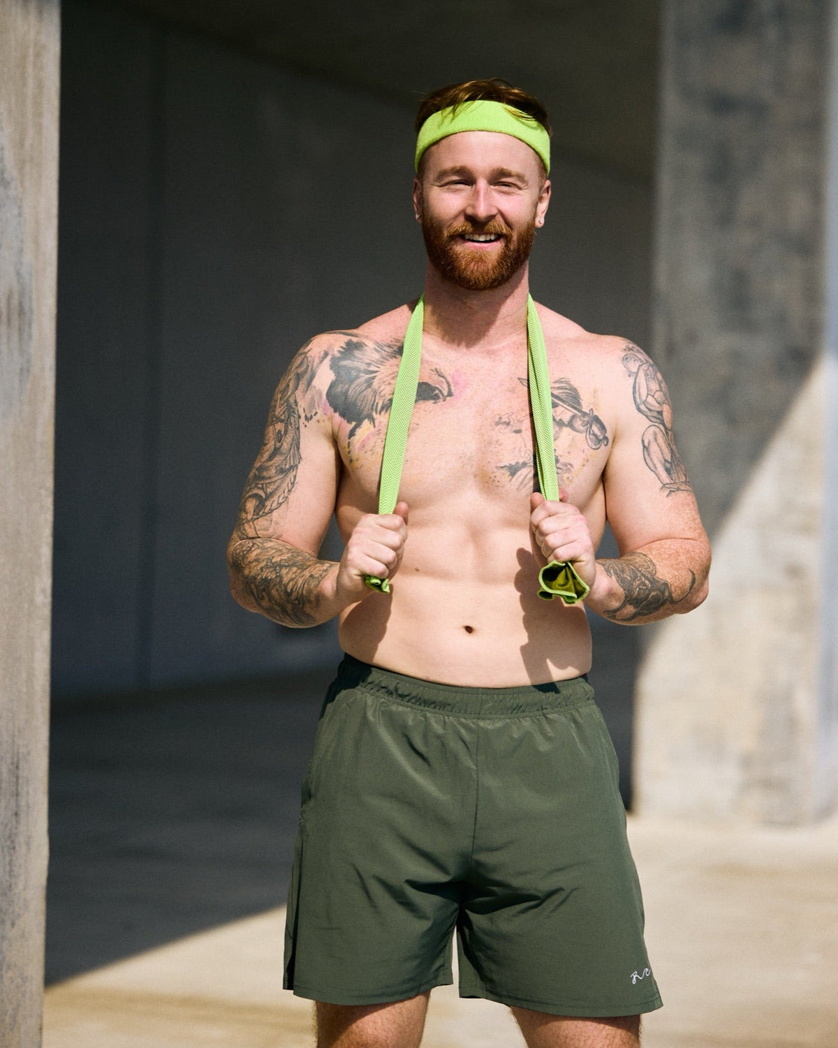 A shirtless man with tattoos stands in a concrete tunnel wearing an olive green athletic headband and matching shorts, holding neon yellow-green resistance bands around his neck.