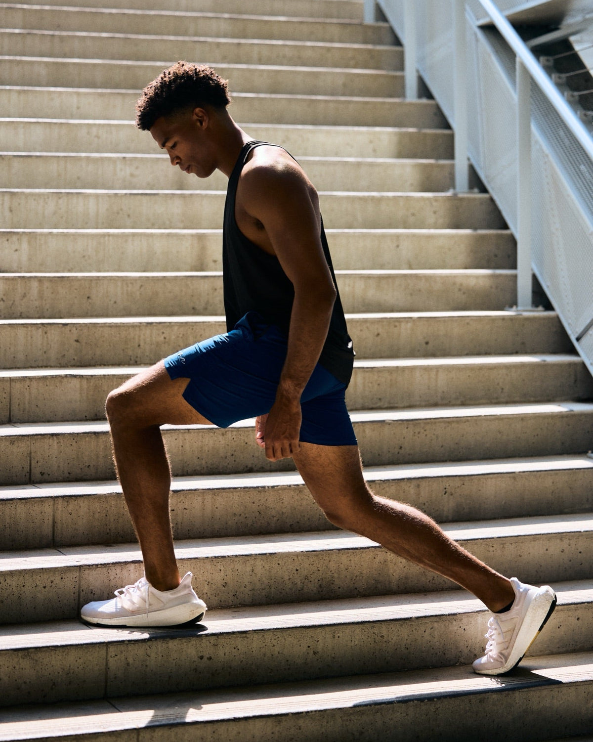 A man in athletic wear, featuring a black tank top, blue shorts, and white sneakers, performing a lunging exercise on outdoor concrete stairs.