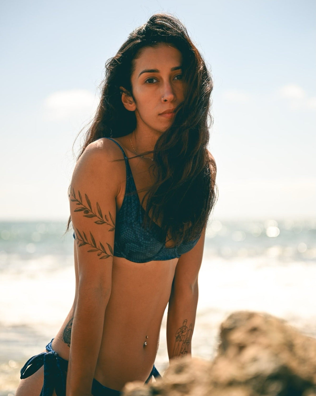 A woman posing on a beach wearing a dark blue bikini, with ocean waves and rocks in the background.