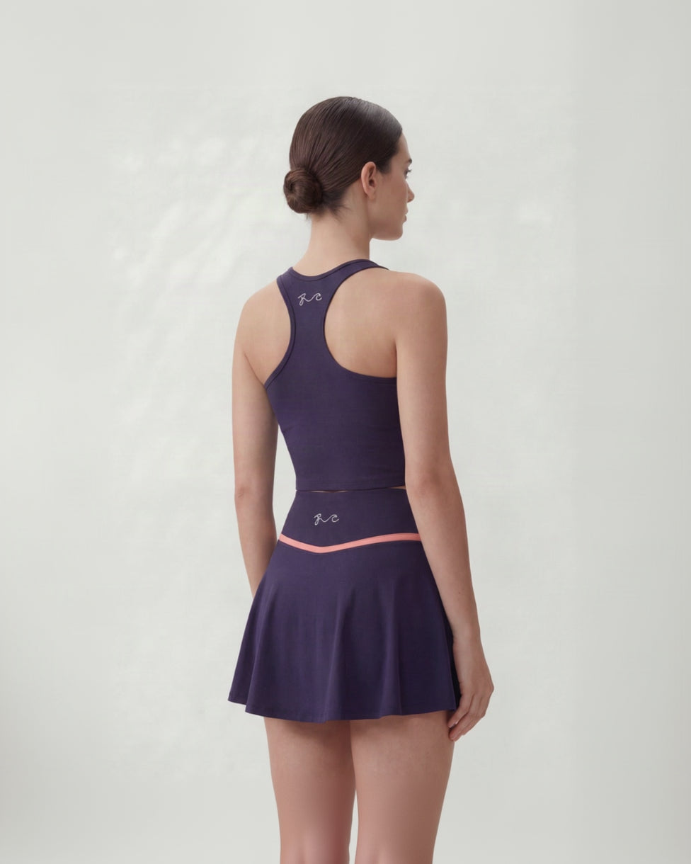 A woman shown from the back wearing a sleeveless athletic dress in deep purple with a thin coral-pink waistband stripe and small white script logos near the neckline and waist.