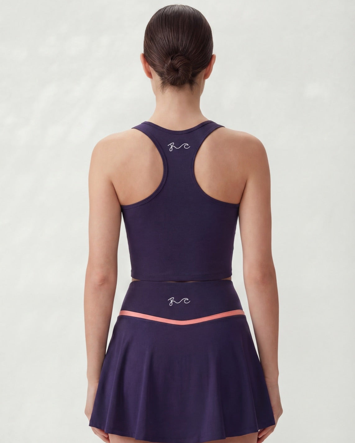 A woman shown from the back wearing a dark purple athletic dress with a flared skirt and a thin coral-pink waistband, against a light gray studio background.