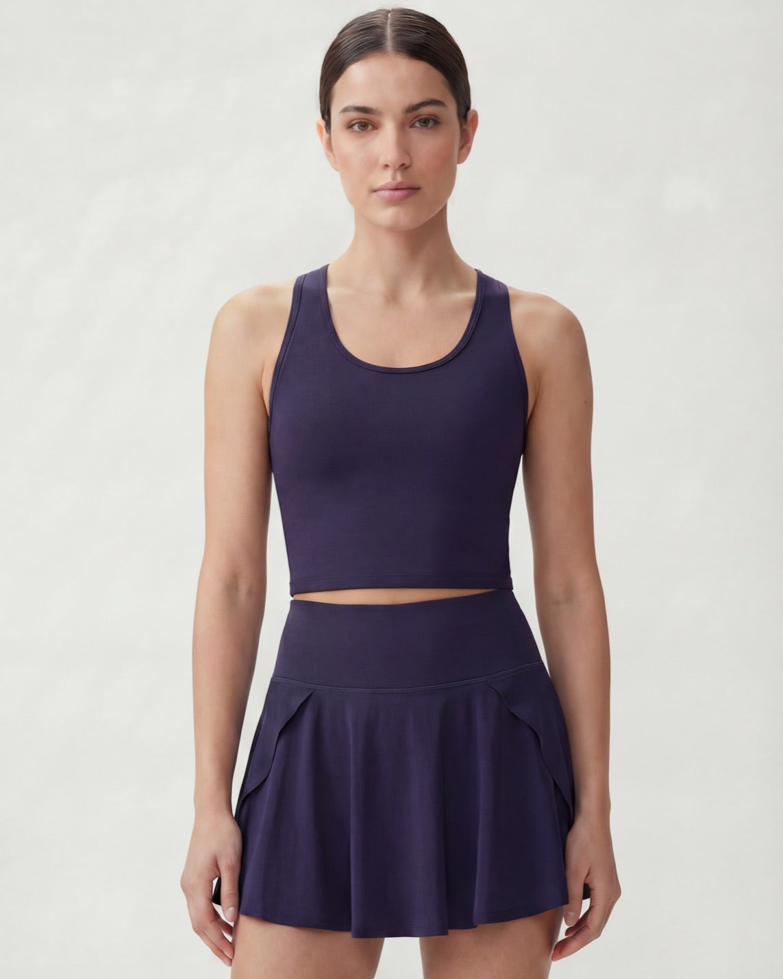 A woman modeling a matching athletic set consisting of a dark purple scoop-neck crop top and a high-waisted pleated mini skirt, photographed against a light gray studio background.