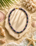 A beaded necklace with purple and dark blue beads and a small gold shell charm, displayed on a stone slab over a sandy beach-themed background with seashells and starfish.