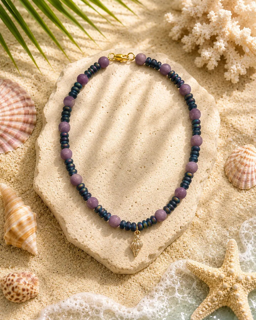 A beaded necklace with purple and dark blue beads and a small gold shell charm, displayed on a stone slab over a sandy beach-themed background with seashells and starfish.