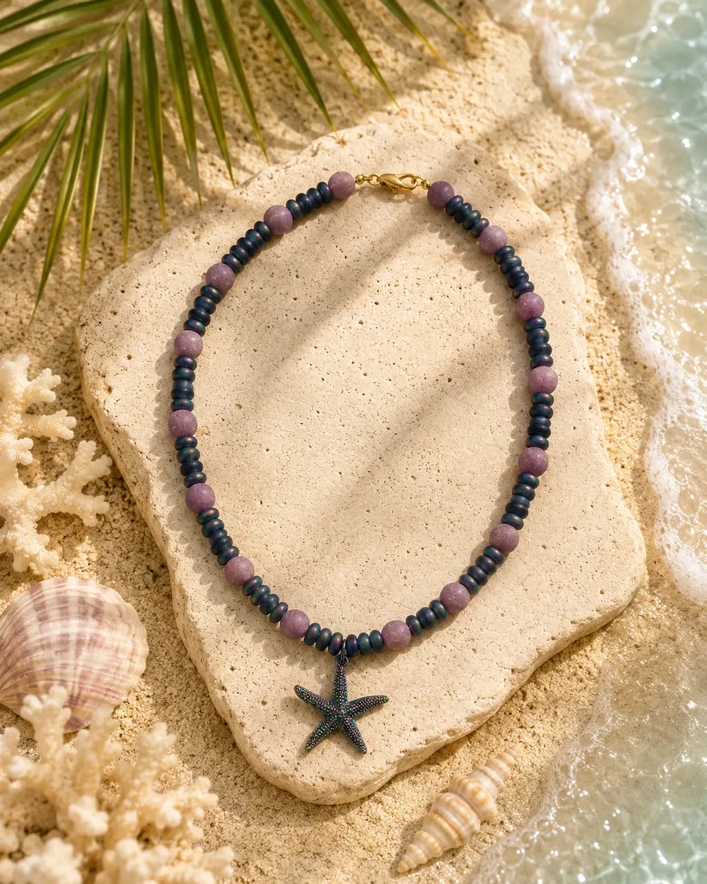 A beaded necklace with dark blue-gray beads, lavender-purple spacer beads, and a dark starfish pendant, styled on a stone over sand with seashells and palm leaves.