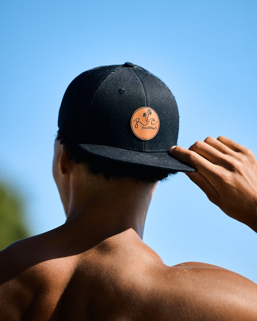 A man viewed from behind wearing a black flat-brim baseball cap with a circular tan leather patch featuring a stylized 'W' logo, set against a clear blue sky.