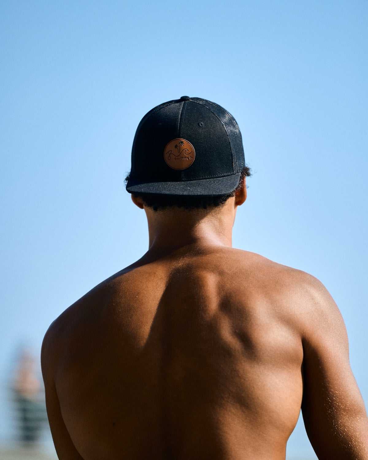 A shirtless man viewed from behind wearing a black baseball cap with a circular brown logo patch, set against a clear blue sky.