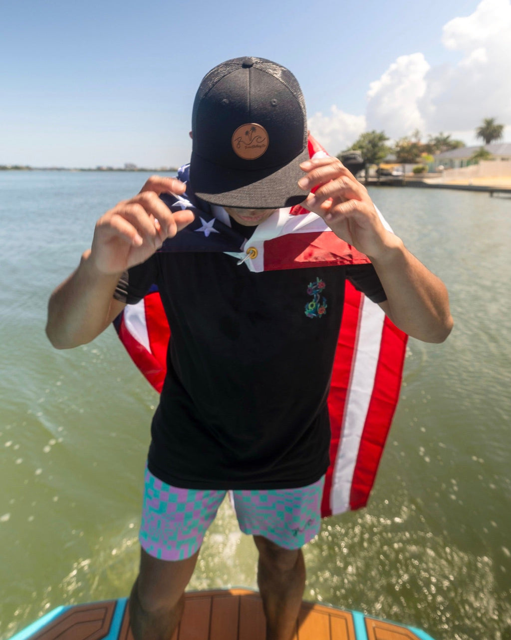 A man standing on a boat wearing a black baseball cap with a brown circular logo, a black t-shirt with a red, white, and blue American flag graphic, and light blue patterned swim trunks, with the flag draped over his shoulders.