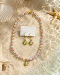A jewelry set featuring a pearl necklace and matching drop earrings with sea turtle charms, styled on a sandy beach background with starfish and a seashell.