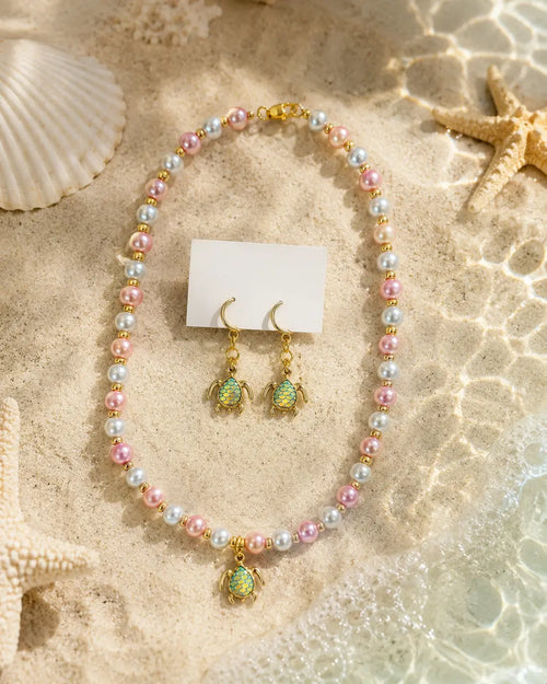 A jewelry set featuring a pearl necklace and matching drop earrings with sea turtle charms, styled on a sandy beach background with starfish and a seashell.