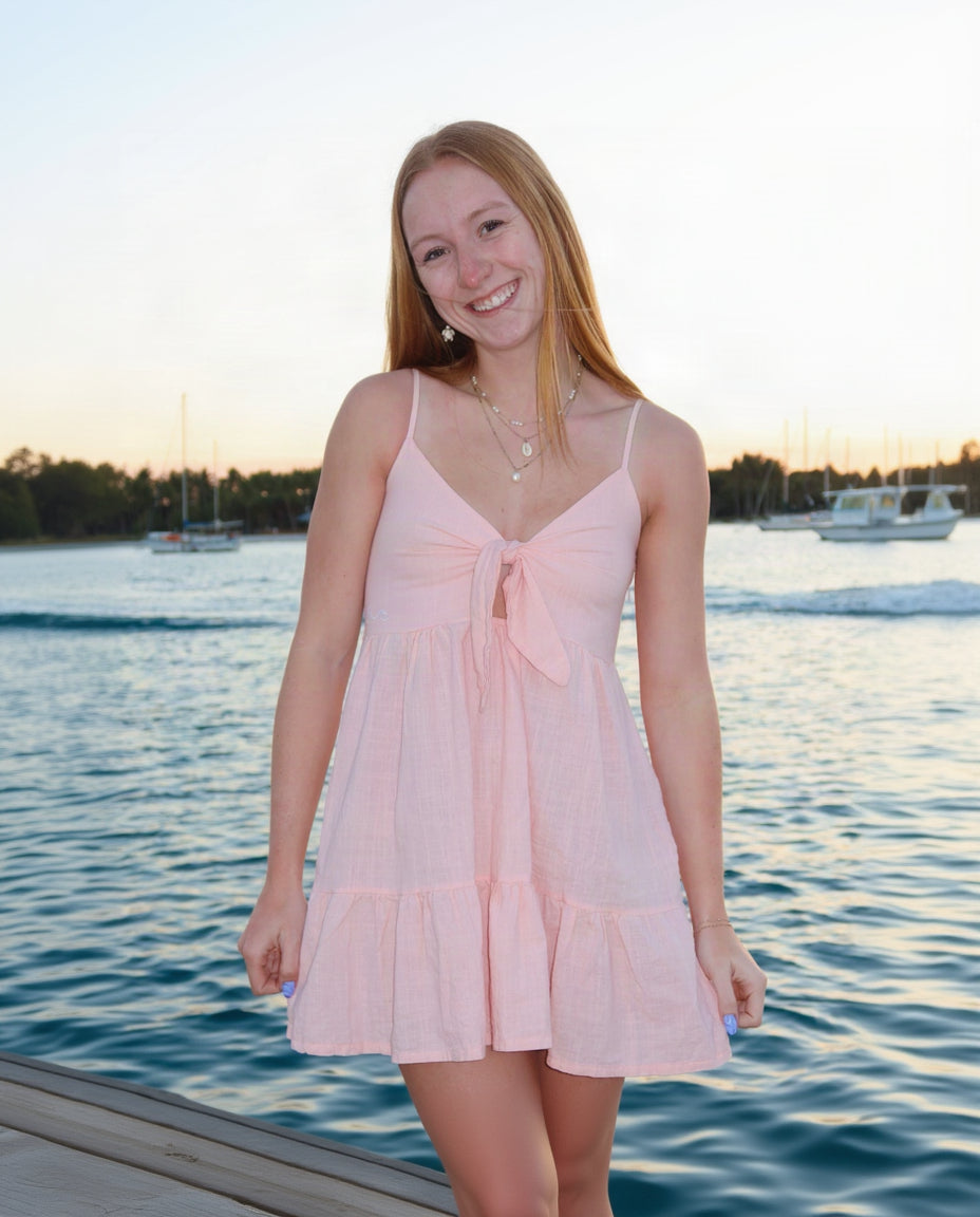 A smiling young woman with long blonde hair stands on a dock by the water wearing a light pink tiered mini dress with thin straps and a front bow detail. Boats and a shoreline are visible in the background at sunset.