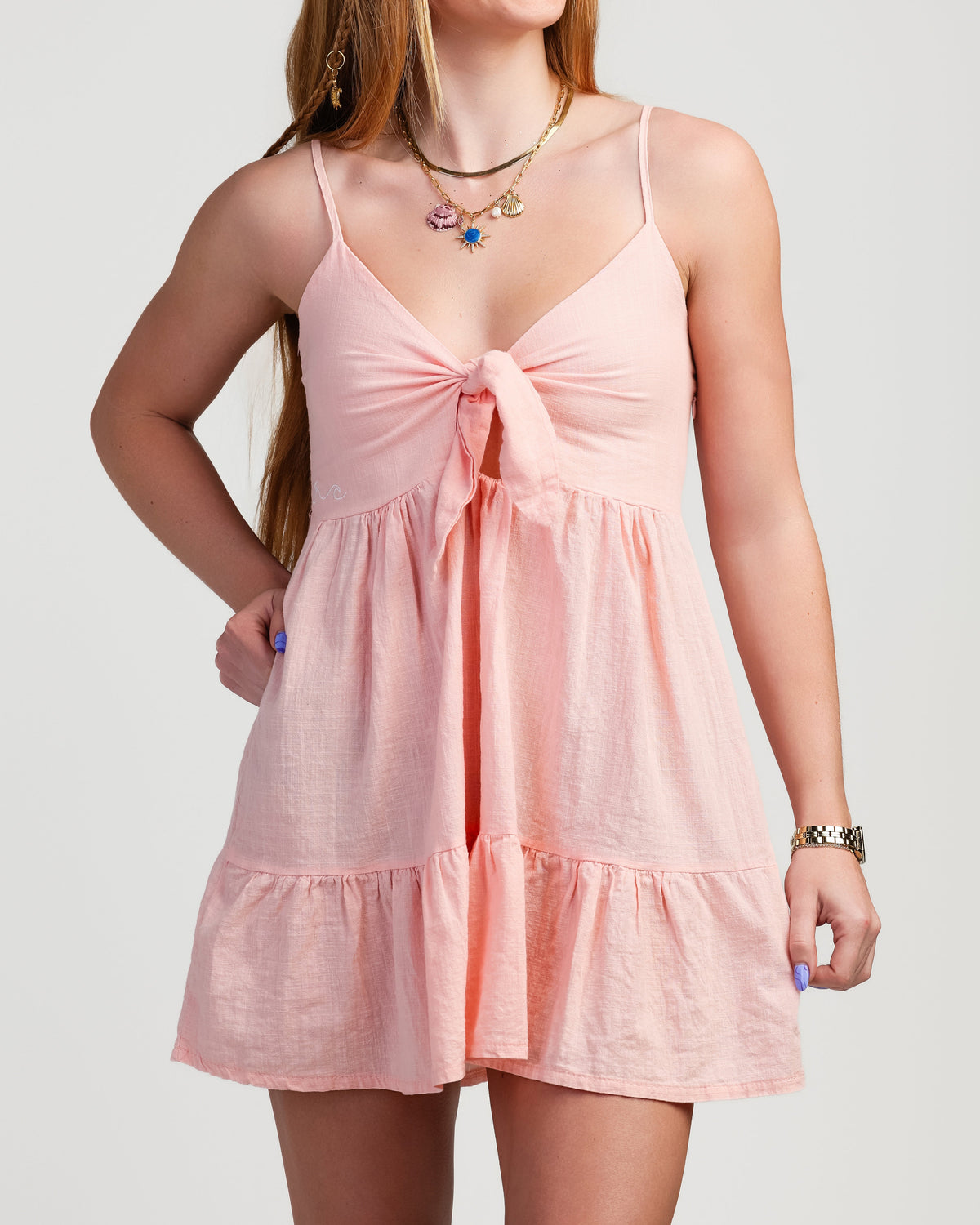 A woman wearing a light pink tiered mini dress with spaghetti straps and a front bow detail, photographed against a plain light gray studio background.