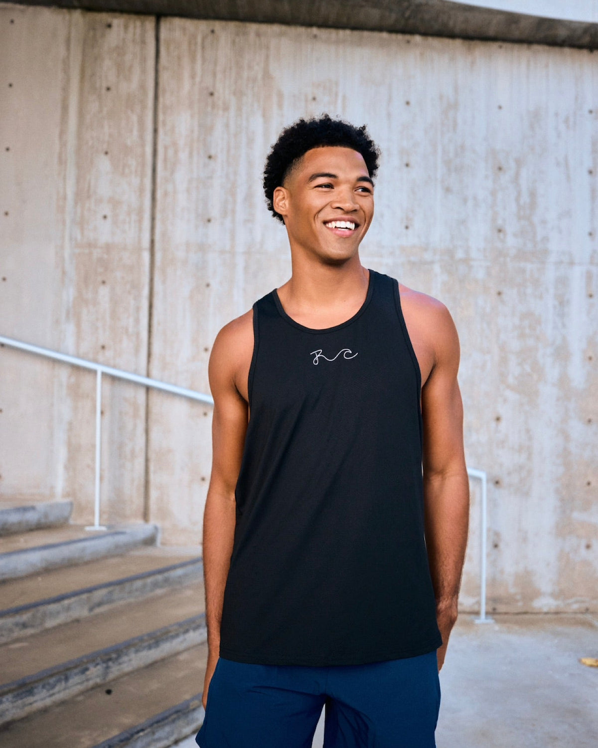A smiling man wearing a black sleeveless athletic tank top with a small white chest logo and navy blue shorts, standing outdoors in front of a concrete wall and steps.