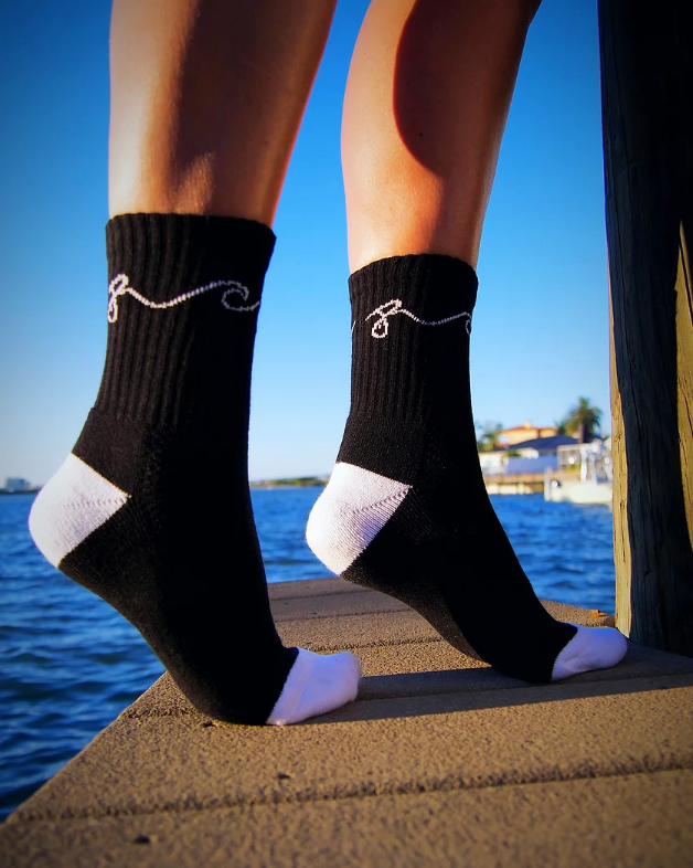 Black ankle socks with white toe and heel sections, featuring a white decorative swirl design near the top, worn on a person's legs standing on a wooden dock with water and a blue sky in the background.
