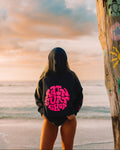 A person standing on a beach facing the ocean, wearing a black hoodie with a bright pink 'RAIN SURF SHOP' graphic on the back. A weathered wooden post with colorful graffiti is visible on the right side of the frame.