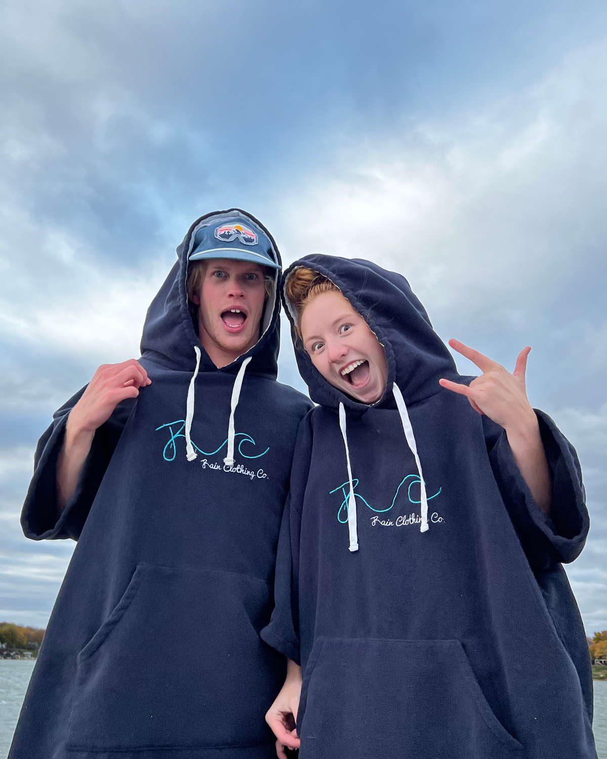 Two young men posing outdoors by a lake under a cloudy sky, wearing matching oversized black hoodies with white drawstrings and a blue graphic cap. The hoodies feature a light blue script logo on the chest.