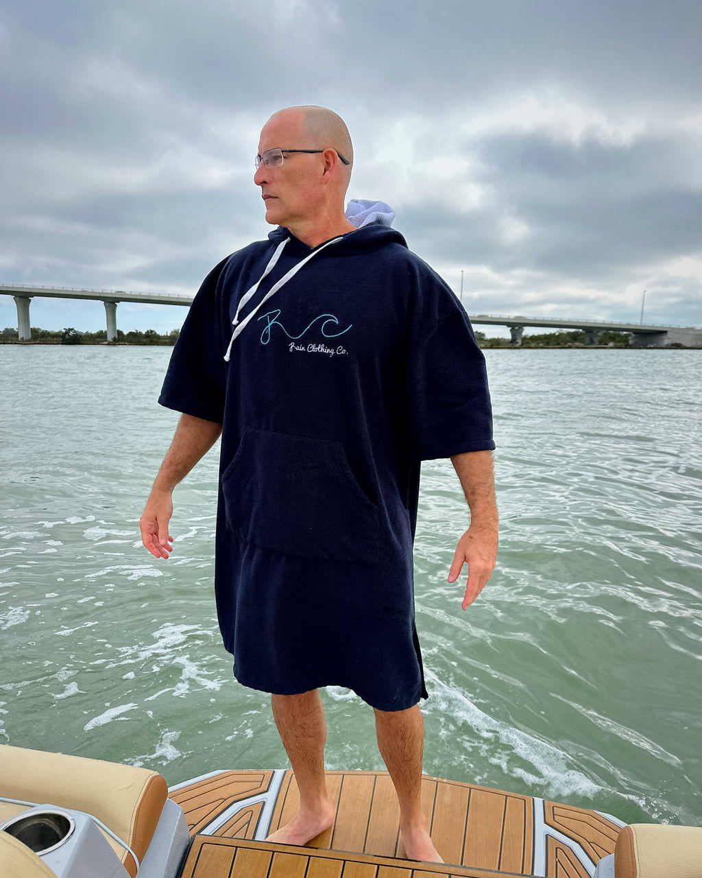 A man wearing a navy blue hooded towel robe stands on the deck of a boat with water and a bridge in the background. The robe has white drawstrings and a light blue inner hood lining, with a white script logo on the chest that reads 'RC Fine Clothing Co.