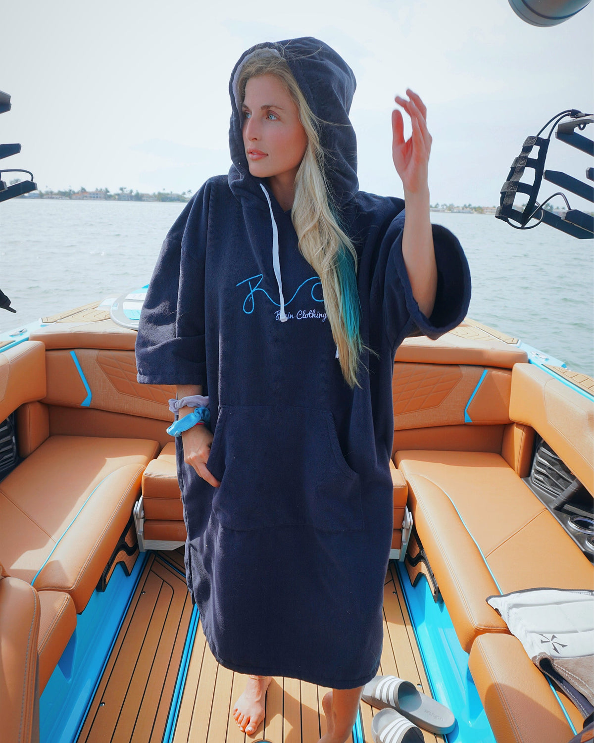 A woman stands on the deck of a boat wearing a navy blue hooded towel dress with a white drawstring and a light blue graphic print on the chest. The background shows the boat's tan and blue seating, wooden deck flooring, and water.