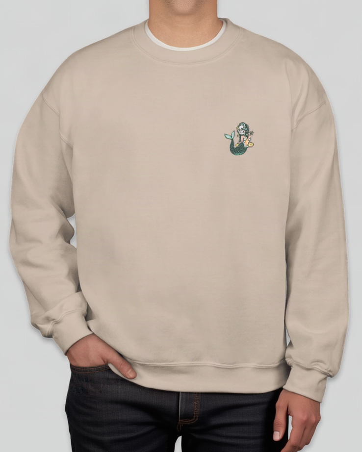 A person wearing a light beige crewneck sweatshirt with a small mermaid graphic on the left chest, paired with dark jeans against a solid gray background.