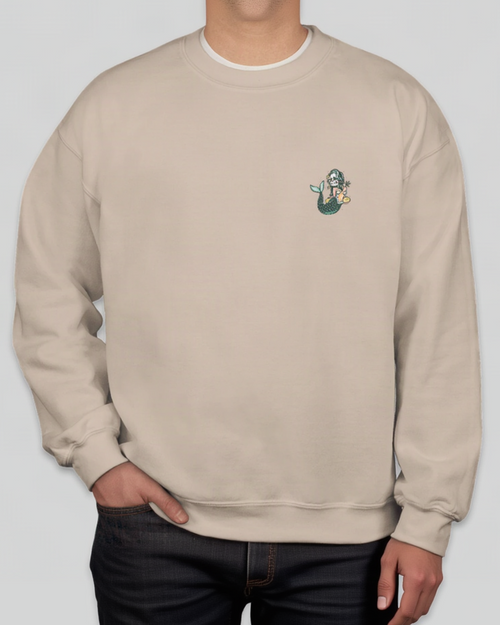 A person wearing a light beige crewneck sweatshirt with a small mermaid graphic on the left chest, paired with dark jeans against a solid gray background.