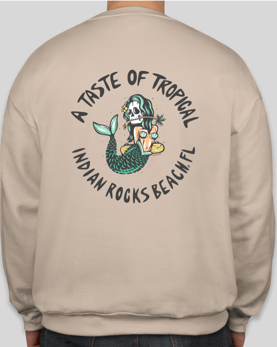 A person wearing a beige crewneck sweatshirt, showing the back of the garment with a large circular graphic print. The graphic features a cartoon mermaid with a skull in her hair, a tropical shell, and the text 'A TASTE OF TROPICAL INDIAN ROCKS BEACH, FL'.