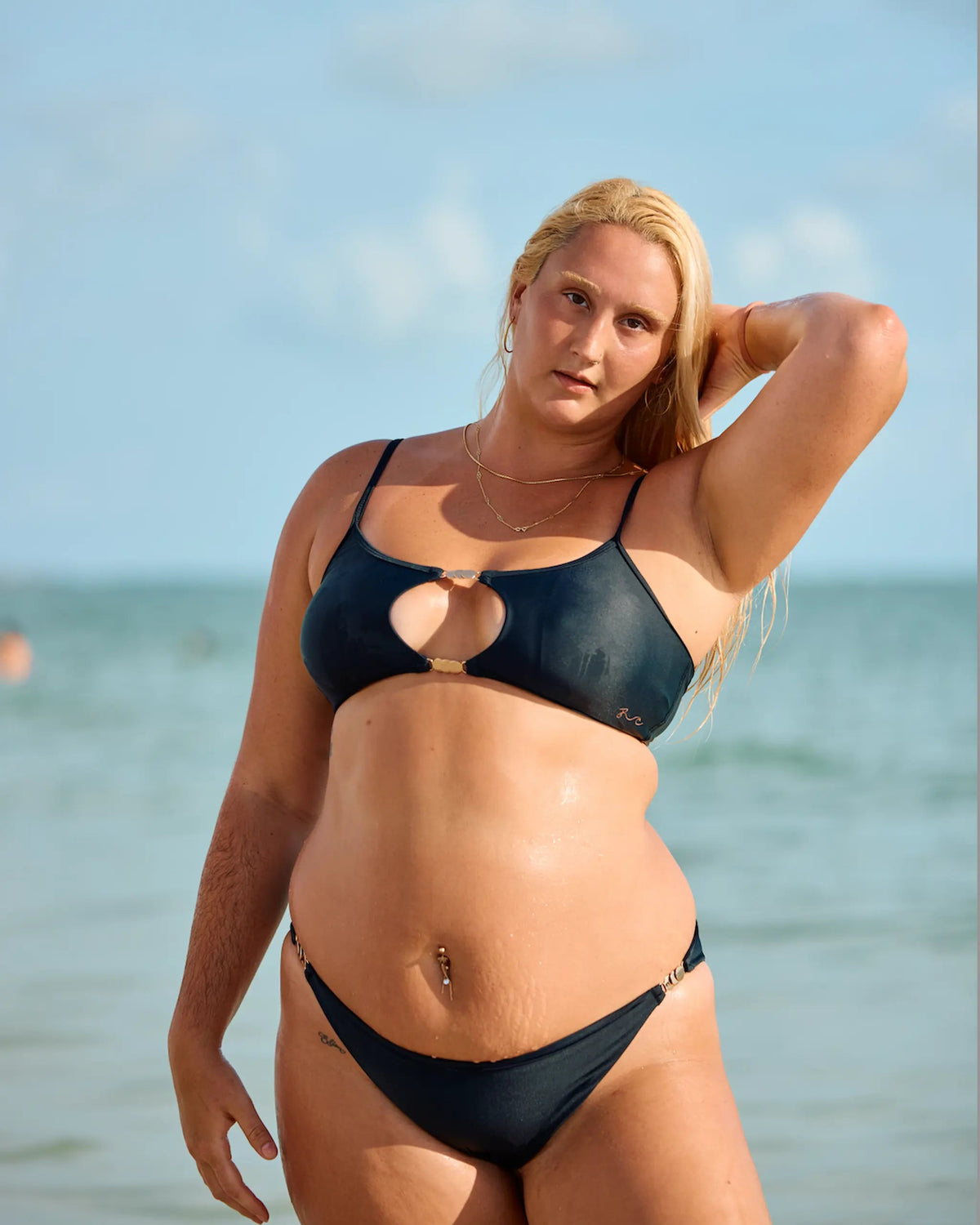 A blonde woman posing on a beach wearing a dark navy blue two-piece bikini with a keyhole cutout on the top.