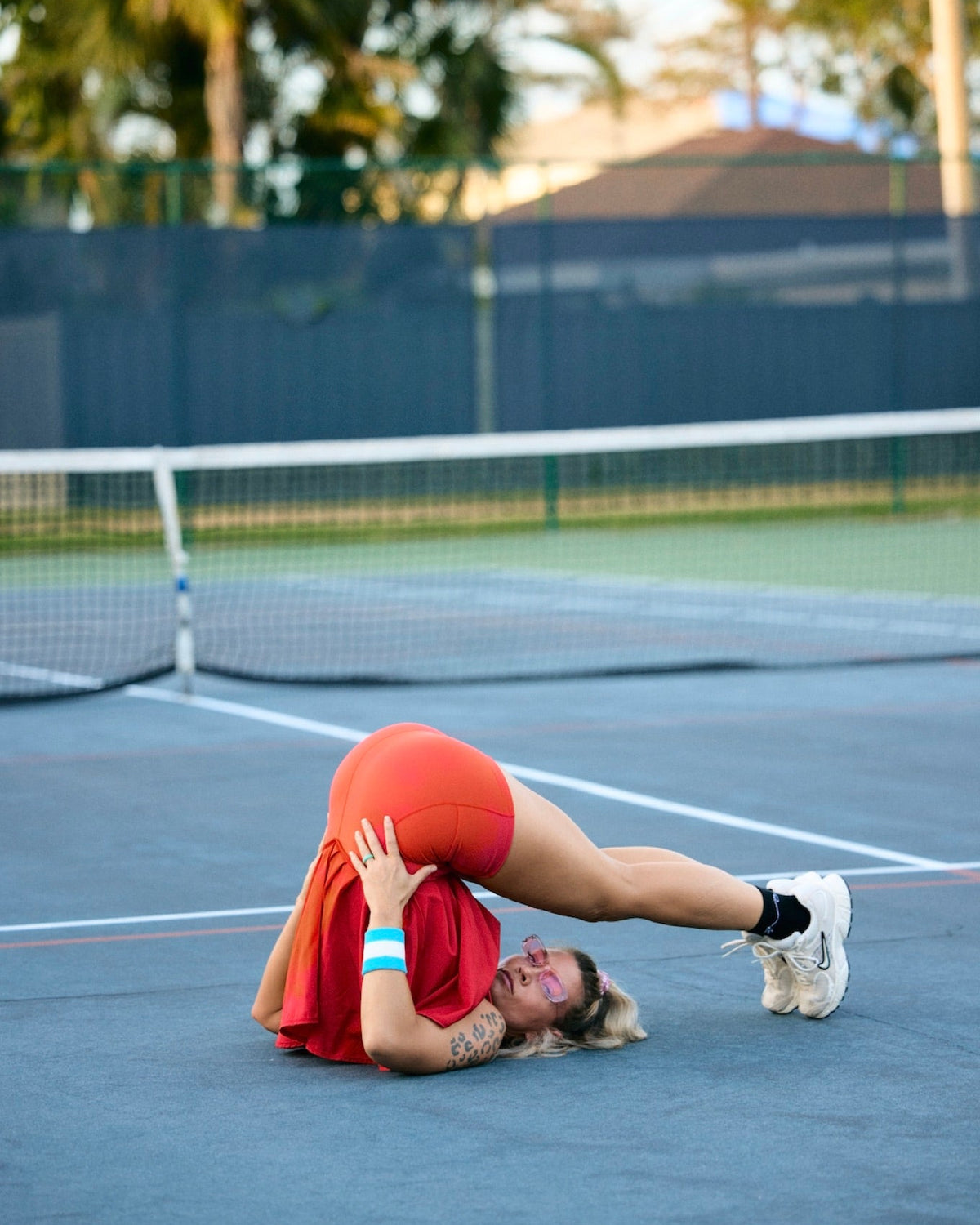 A woman in a red tennis dress performs a dramatic backbend on a blue tennis court, with a net, fence, and palm trees in the background.