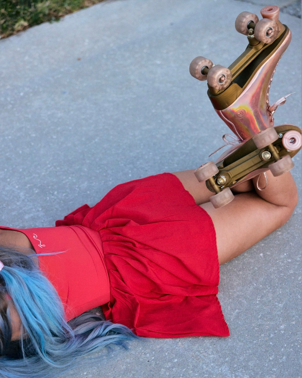 A person lying on gray pavement with one roller skate raised toward the camera. The person is wearing a bright red dress and has blue-to-brown hair.