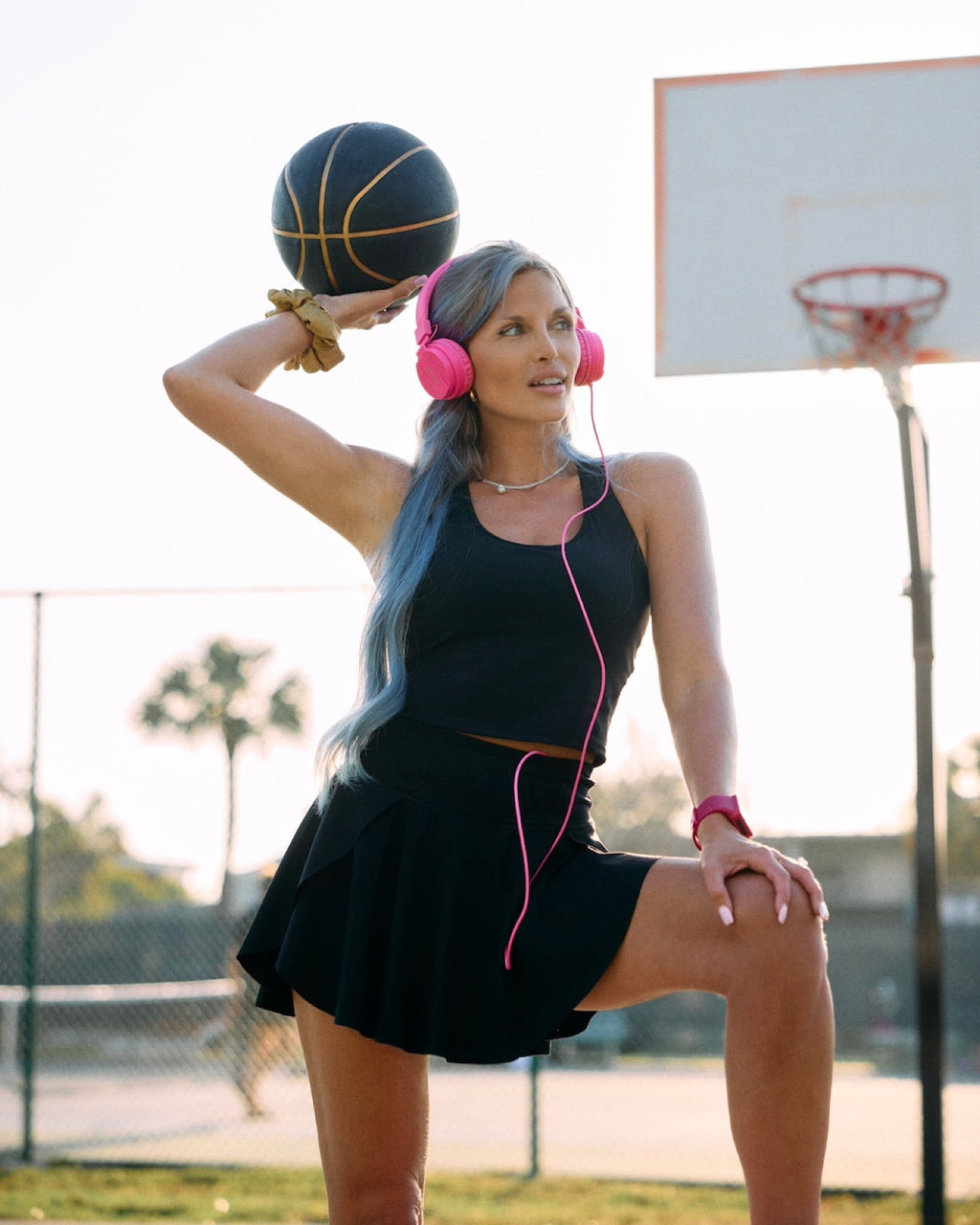 A woman with blue hair poses on an outdoor basketball court wearing a black athletic dress, pink headphones, and pink earphones while holding a basketball.