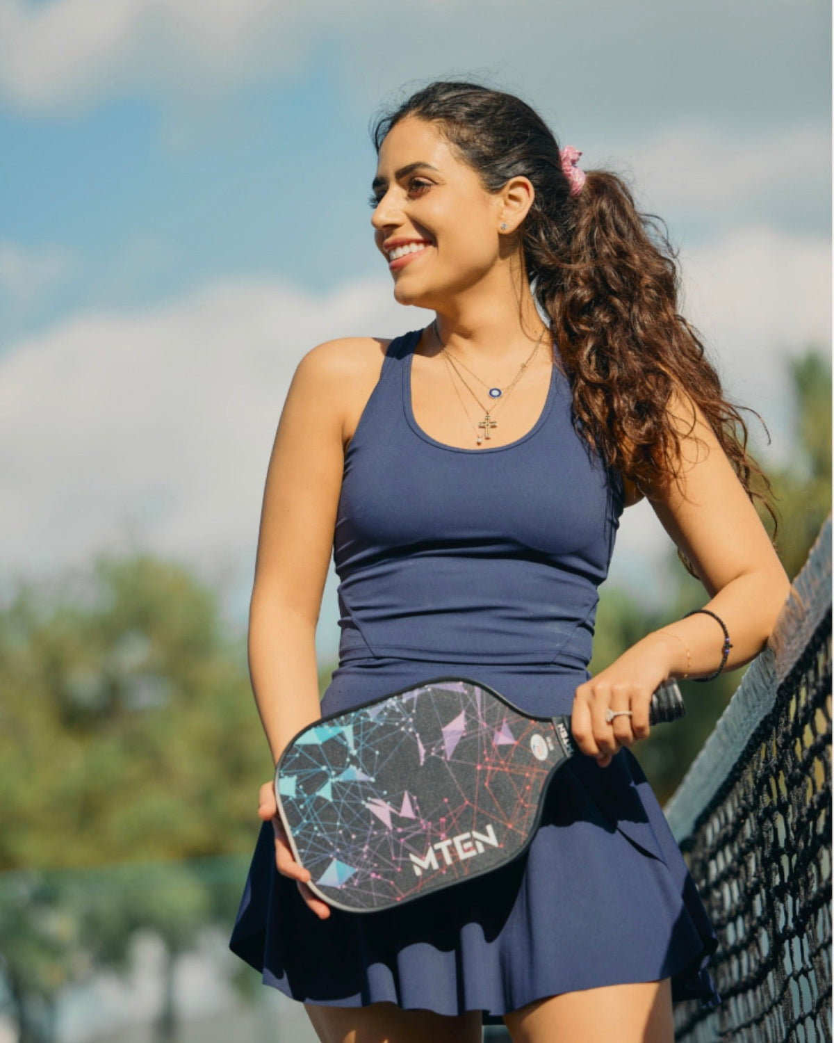 A woman on a pickleball court wearing a navy blue tank top and matching skirt, holding a pickleball paddle with the brand name 'Miten' visible. The background shows a sunny outdoor court setting with sky, clouds, and netting.