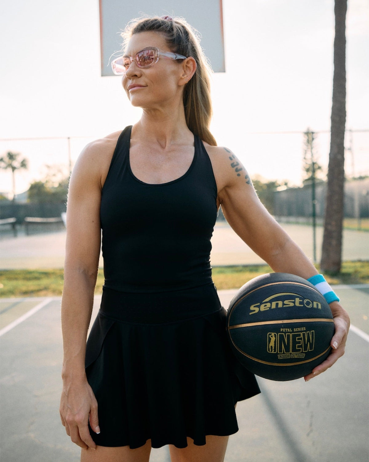 A woman wearing a black athletic tank top and matching black skirt stands on an outdoor basketball court holding a Sanston basketball.