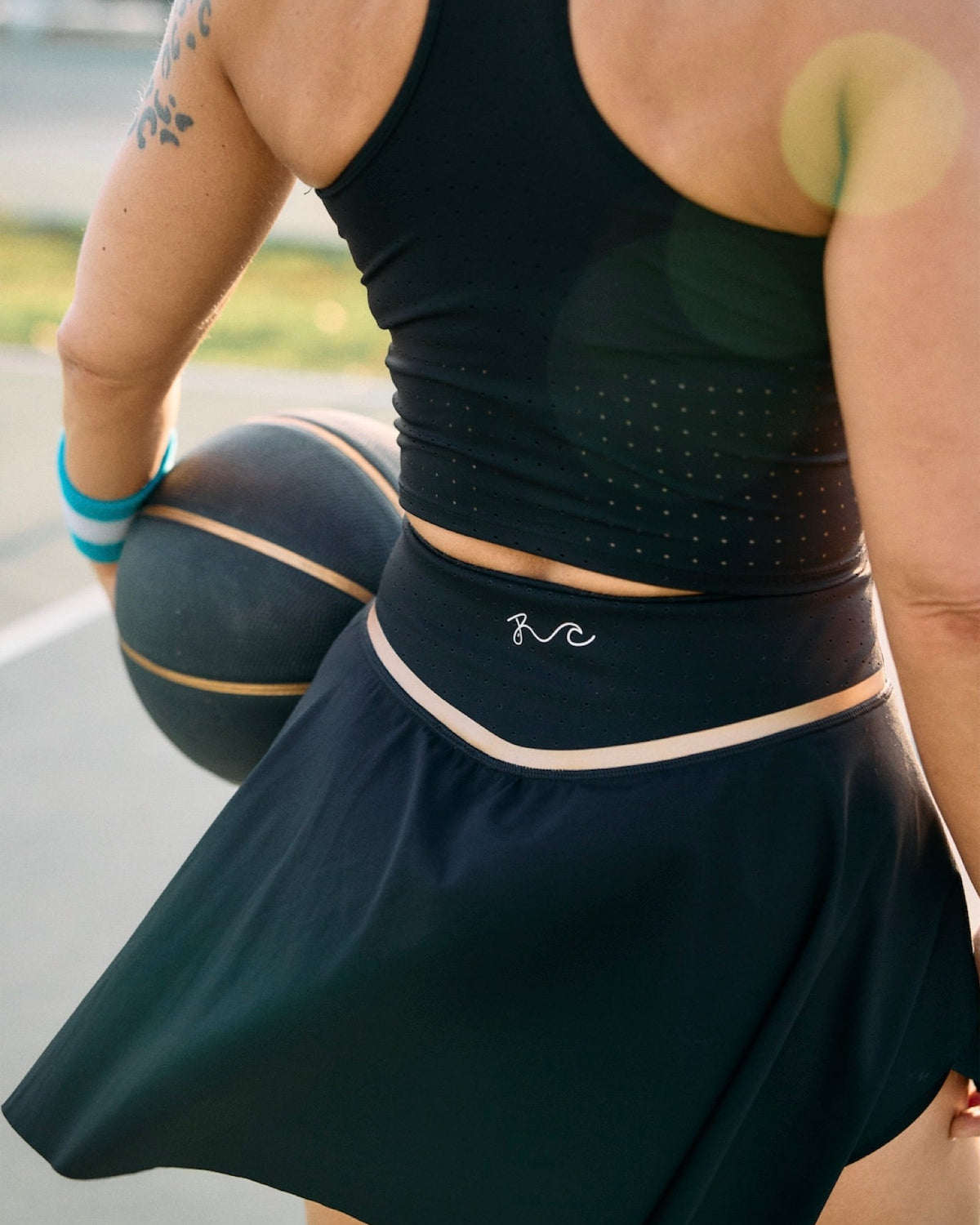 Rear view of a woman on an outdoor basketball court wearing a black athletic tank top and matching flared skirt with a thin beige stripe detail at the waistband, holding a basketball.